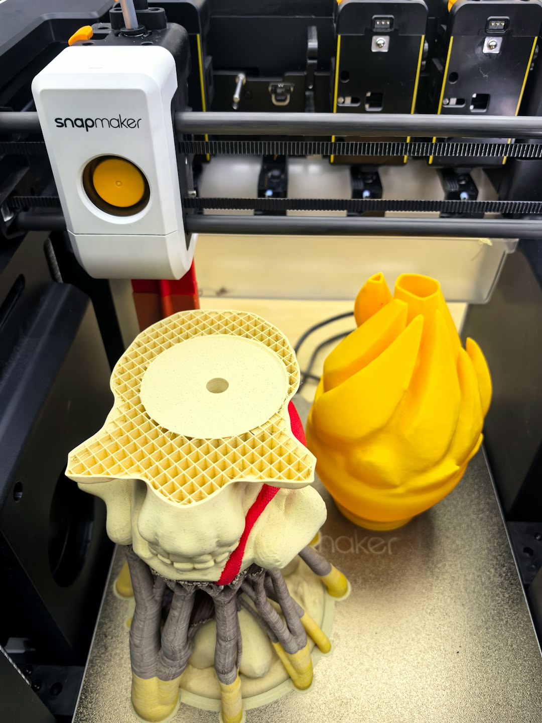 Stampante 3D crea una creatura beige e grigia e una scultura astratta gialla.